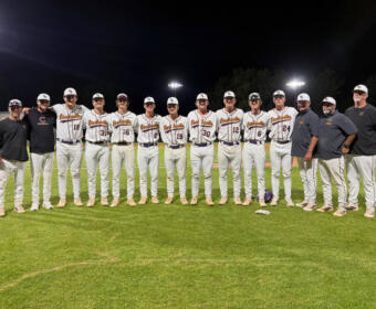 A baseball team stands in a line on the field at night for a group photo, wearing white uniforms with maroon writing and caps.