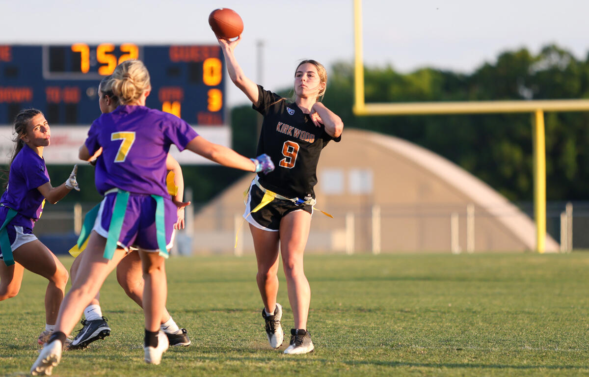 Female flag football player in a black jersey (number 9) throwing a pass, with teammates in purple nearby on a grassy field.
