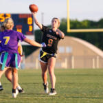 Female flag football player in a black jersey (number 9) throwing a pass, with teammates in purple nearby on a grassy field.