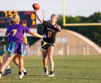 Female flag football player in a black jersey (number 9) throwing a pass, with teammates in purple nearby on a grassy field.