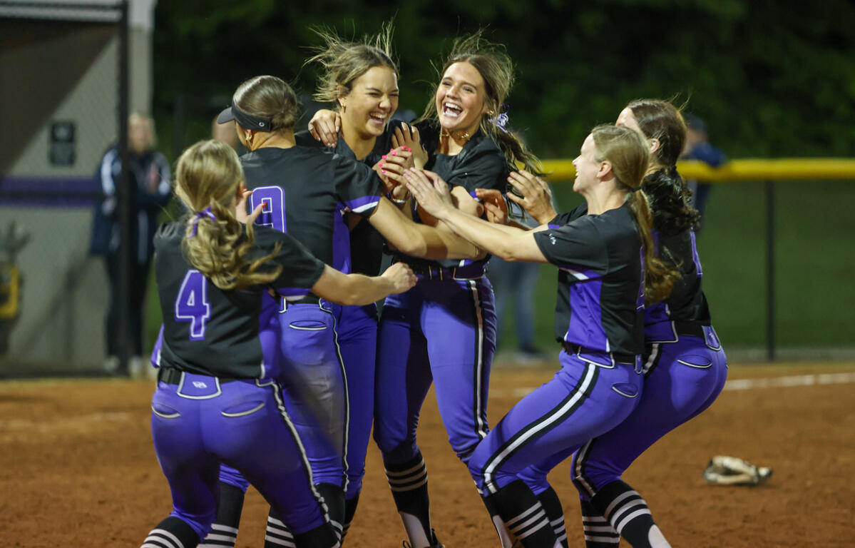 A women's softball team in purple and black uniforms celebrates, hugging and cheering on the infield dirt at night.