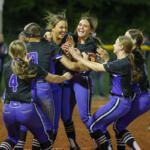 A women's softball team in purple and black uniforms celebrates, hugging and cheering on the infield dirt at night.