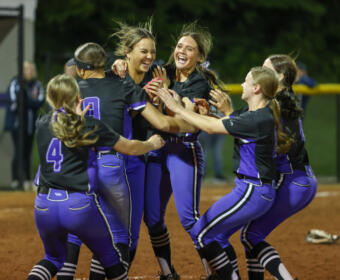 A women's softball team in purple and black uniforms celebrates, hugging and cheering on the infield dirt at night.