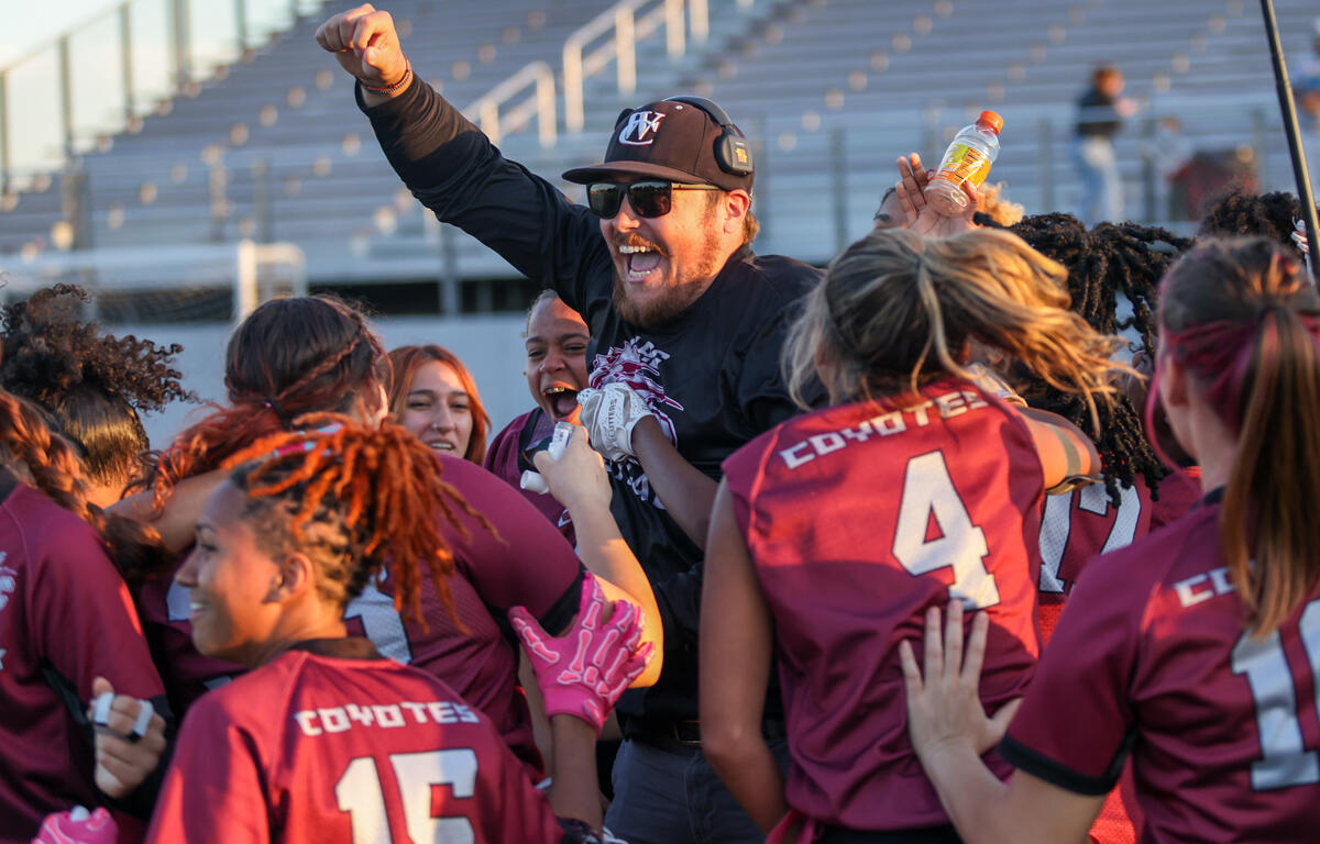 Coach being lifted by a cheering girls' team in maroon uniforms on a stadium field, celebrating a win.