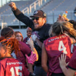 Coach being lifted by a cheering girls' team in maroon uniforms on a stadium field, celebrating a win.