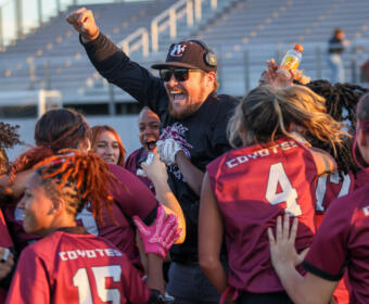 Coach being lifted by a cheering girls' team in maroon uniforms on a stadium field, celebrating a win.