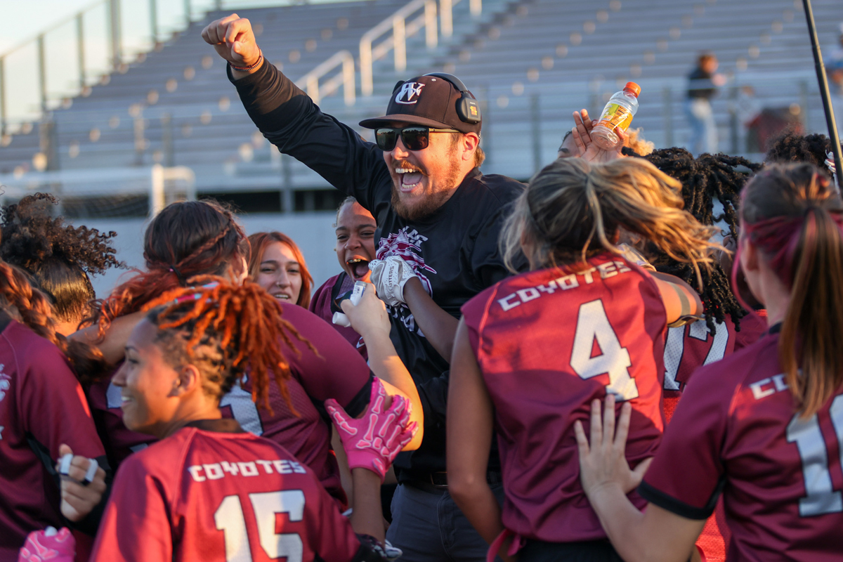 Coach being lifted by a cheering girls' team in maroon uniforms on a stadium field, celebrating a win.