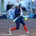 Female softball batter in navy uniform and white helmet swings at a pitched ball on a dirt field, spectators behind a chain-link fence in the stands.