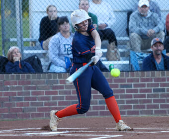 Female softball batter in navy uniform and white helmet swings at a pitched ball on a dirt field, spectators behind a chain-link fence in the stands.