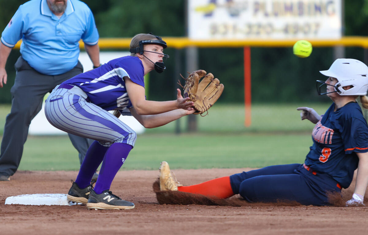 Softball fielder in purple reaches for a ground ball as a runner slides into base, derby-style play on a dirt infield with an umpire nearby.