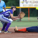 Softball fielder in purple reaches for a ground ball as a runner slides into base, derby-style play on a dirt infield with an umpire nearby.