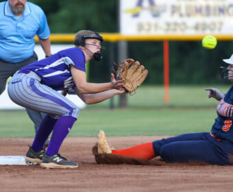Softball fielder in purple reaches for a ground ball as a runner slides into base, derby-style play on a dirt infield with an umpire nearby.