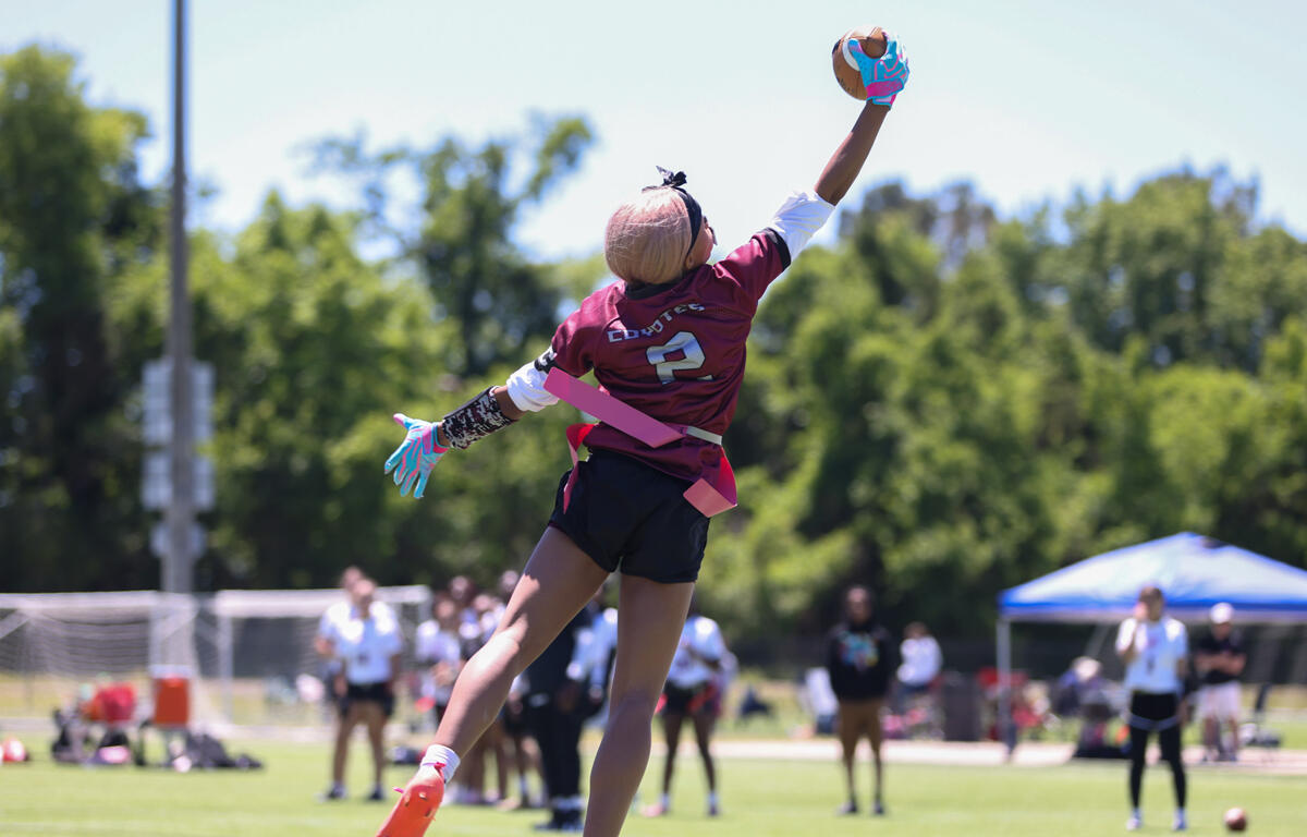Female goalkeeper in maroon jersey #2 leaps to catch a ball with gloved hand during an outdoor field game on a sunny day, with spectators and a blue tent in the background.