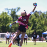 Female goalkeeper in maroon jersey #2 leaps to catch a ball with gloved hand during an outdoor field game on a sunny day, with spectators and a blue tent in the background.