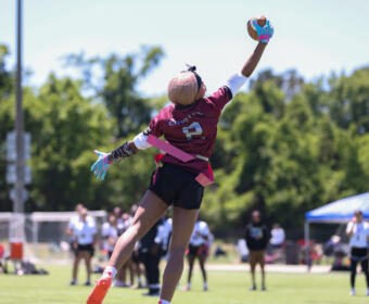 Female goalkeeper in maroon jersey #2 leaps to catch a ball with gloved hand during an outdoor field game on a sunny day, with spectators and a blue tent in the background.