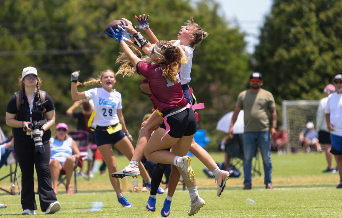 Two female flag football players leap to catch a ball while others watch on a sunny field, a photographer nearby.