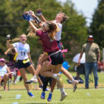 Two female flag football players leap to catch a ball while others watch on a sunny field, a photographer nearby.
