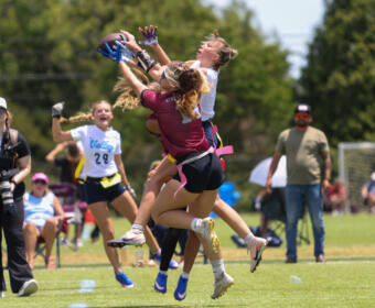 Two female flag football players leap to catch a ball while others watch on a sunny field, a photographer nearby.