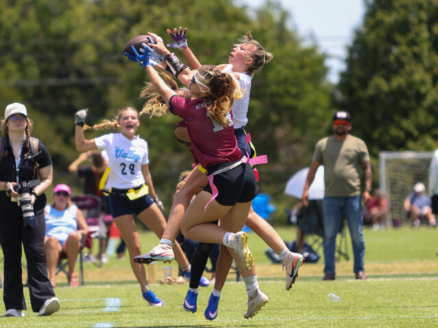 Two female flag football players leap to catch a ball while others watch on a sunny field, a photographer nearby.