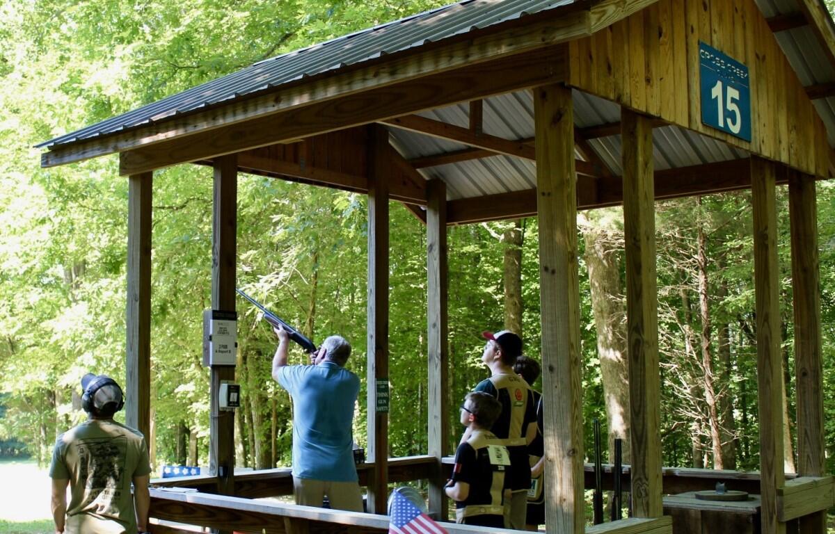 Wooden outdoor shooting range pavilion in a forest; a man fires a shotgun while others watch.