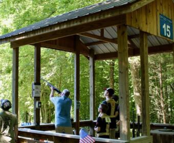 Wooden outdoor shooting range pavilion in a forest; a man fires a shotgun while others watch.