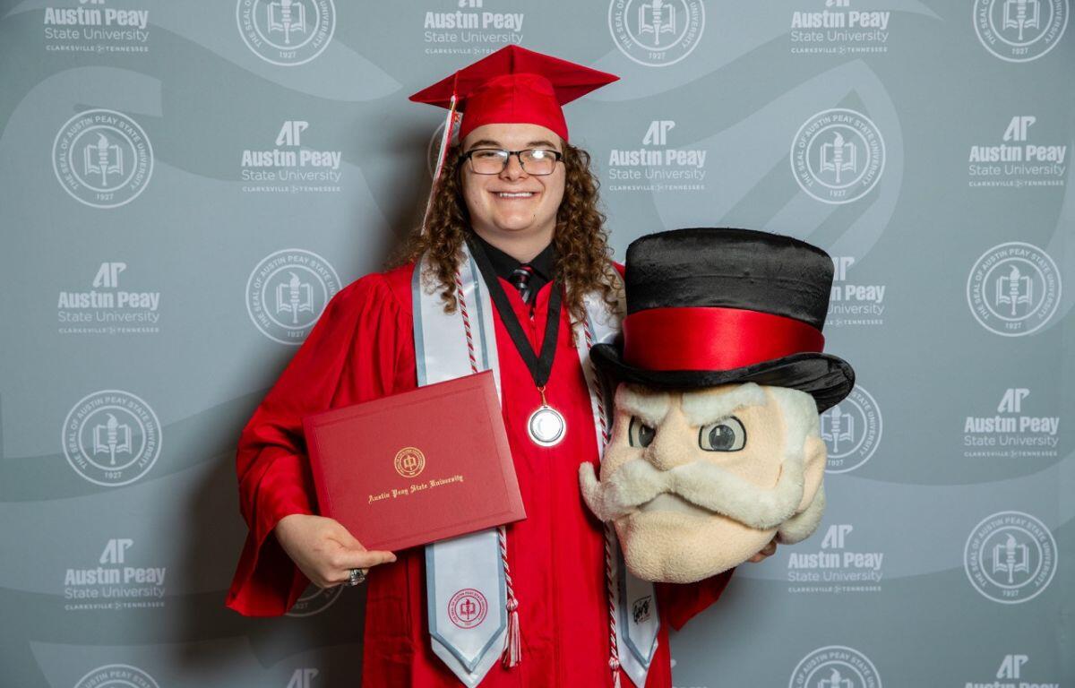 Graduate in red cap and gown holding a diploma folder and a large plush mascot head, against an Austin Peay backdrop.