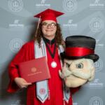Graduate in red cap and gown holding a diploma folder and a large plush mascot head, against an Austin Peay backdrop.