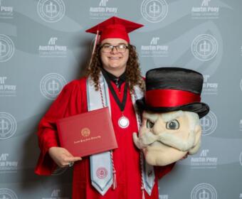 Graduate in red cap and gown holding a diploma folder and a large plush mascot head, against an Austin Peay backdrop.