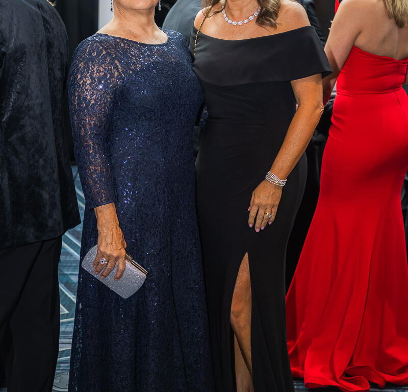 Two women in formal gowns pose for a photo at a gala; one in navy lace with a silver clutch, the other in a black off-the-shoulder gown with a high slit and jewelry.