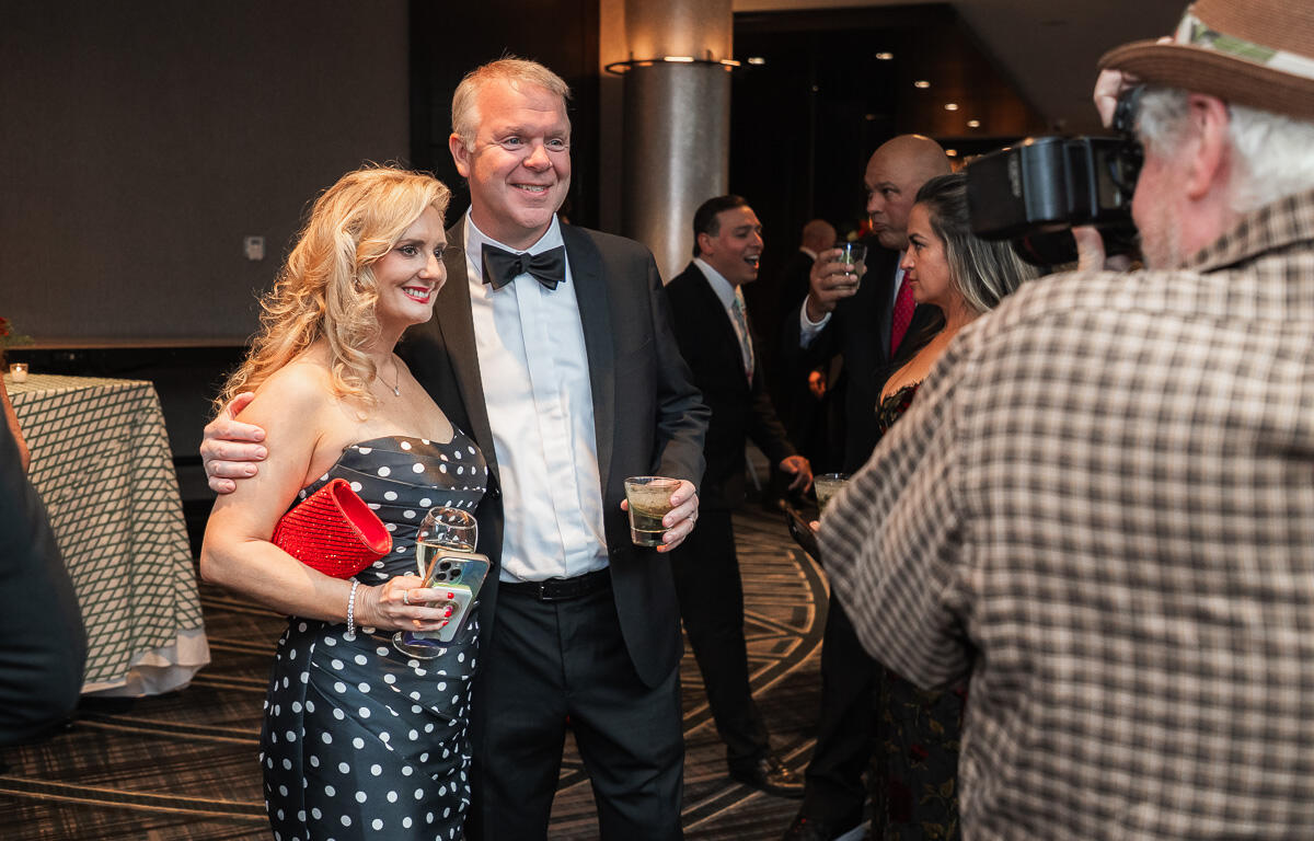 Couple posing at a formal event: woman in polka-dot gown with red clutch and drinks, man in tuxedo with bow tie smiling for a photographer nearby.