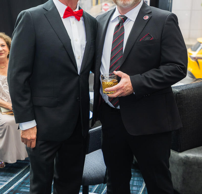 Two men in black suits posing for a photo at a formal event; one wears a red bow tie and the other holds a drink and smiles.