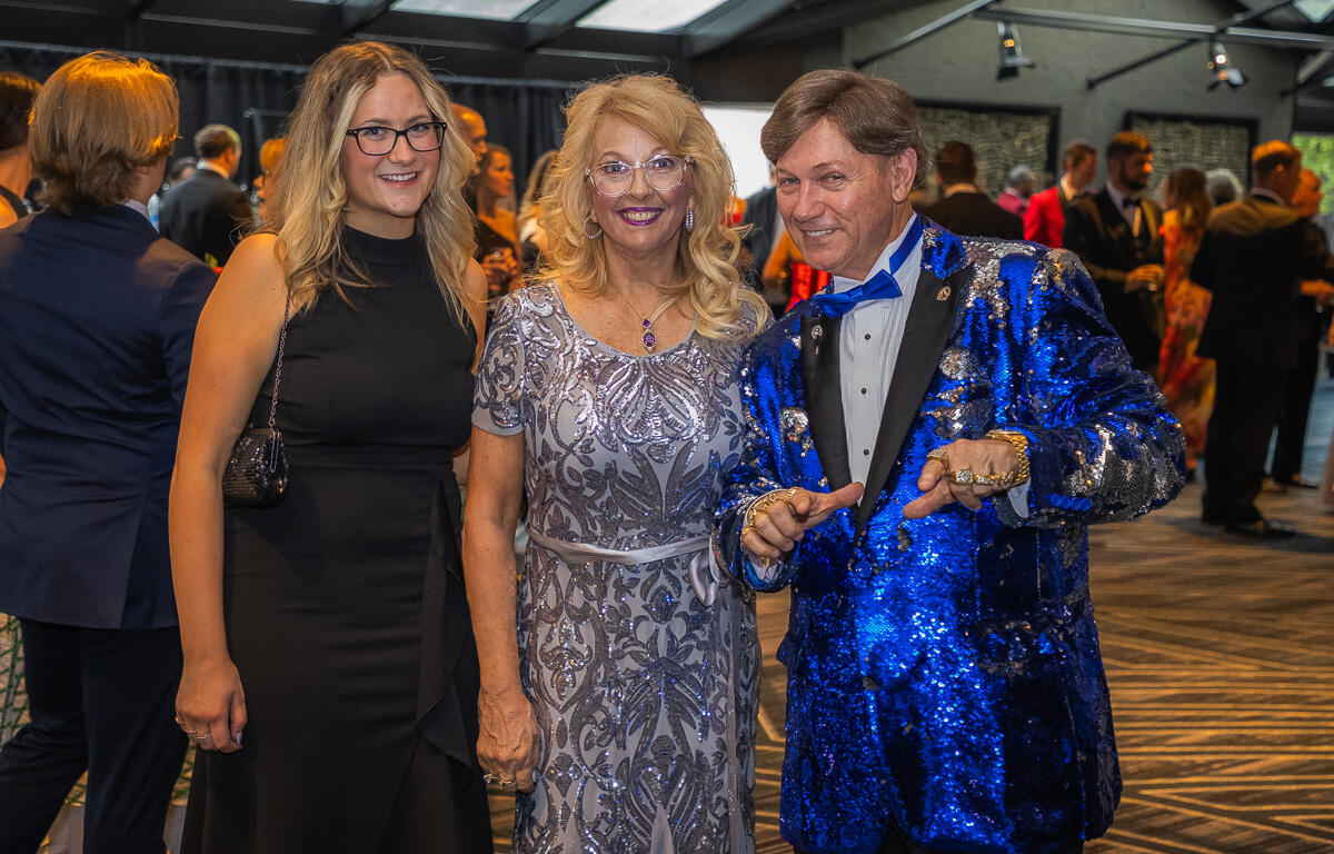 Three adults posing for a photo at a formal event: woman in a black dress, woman in a silver gown, and a man in a blue sequined jacket smile at the camera.