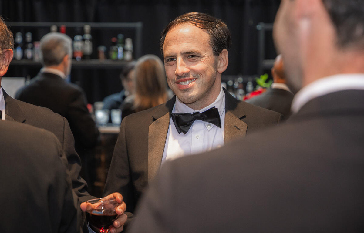 Man in a black tuxedo and bow tie smiles while holding a glass of red wine at a formal cocktail party, with a bar in the background.