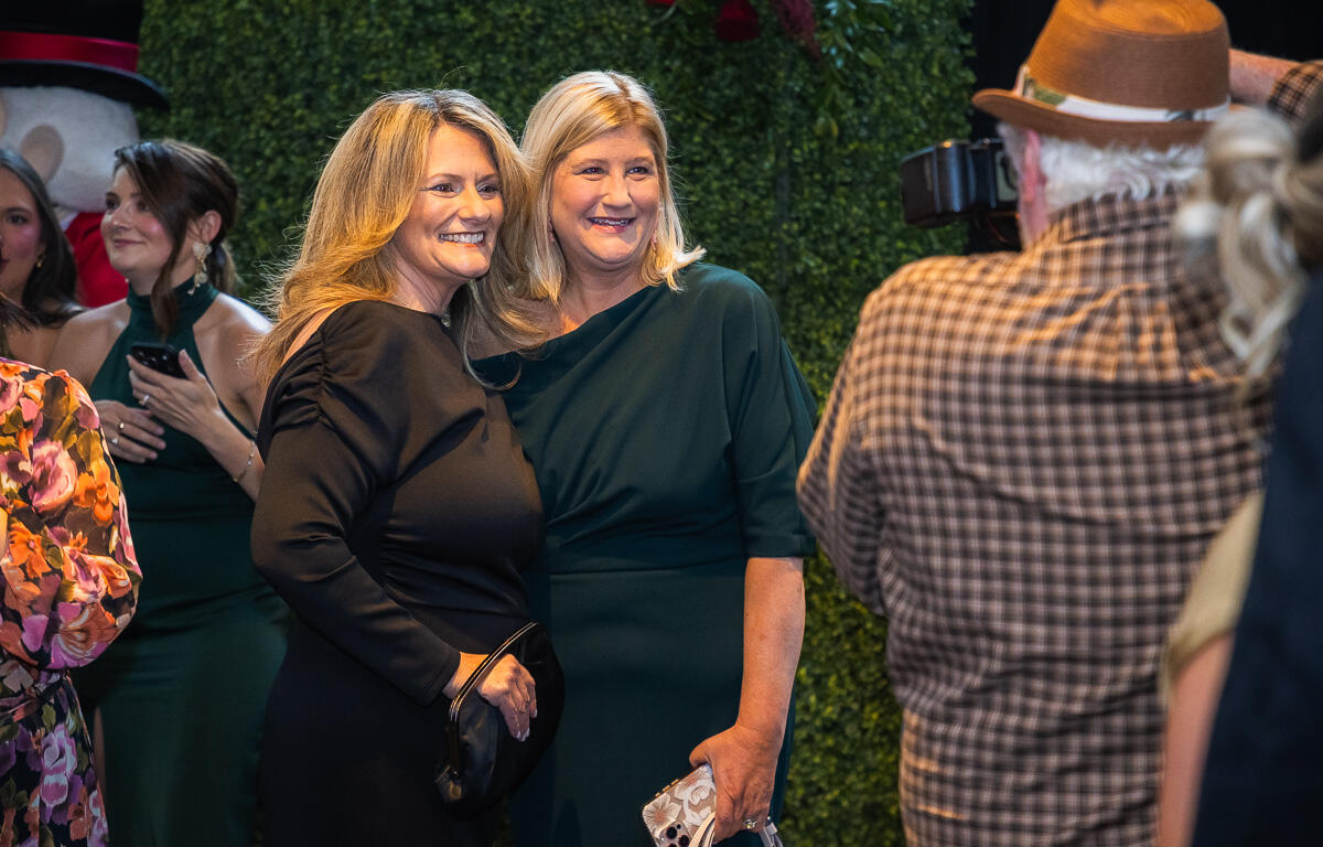 Two smiling women pose for a photo at a formal event, one in black holding a small clutch, the other in dark green dress with a patterned backdrop behind them.