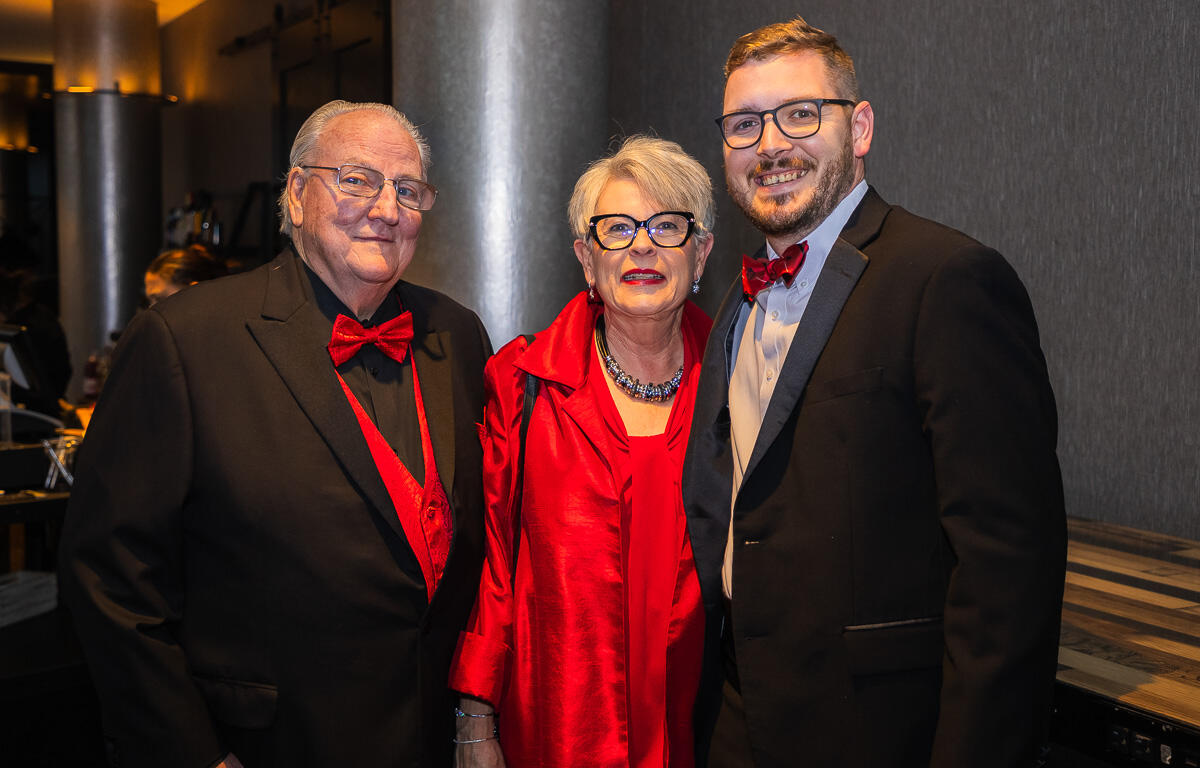Three well-dressed guests posing for a photo at a formal event: two men in tuxedos with red bow ties and a woman in a red dress in the center.
