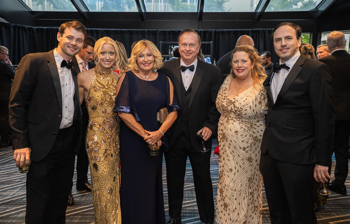 Six adults in formal wear pose together at a black-tie gala, smiling for a group photo English-style venue backdrop behind them so the scene reads as a formal event.