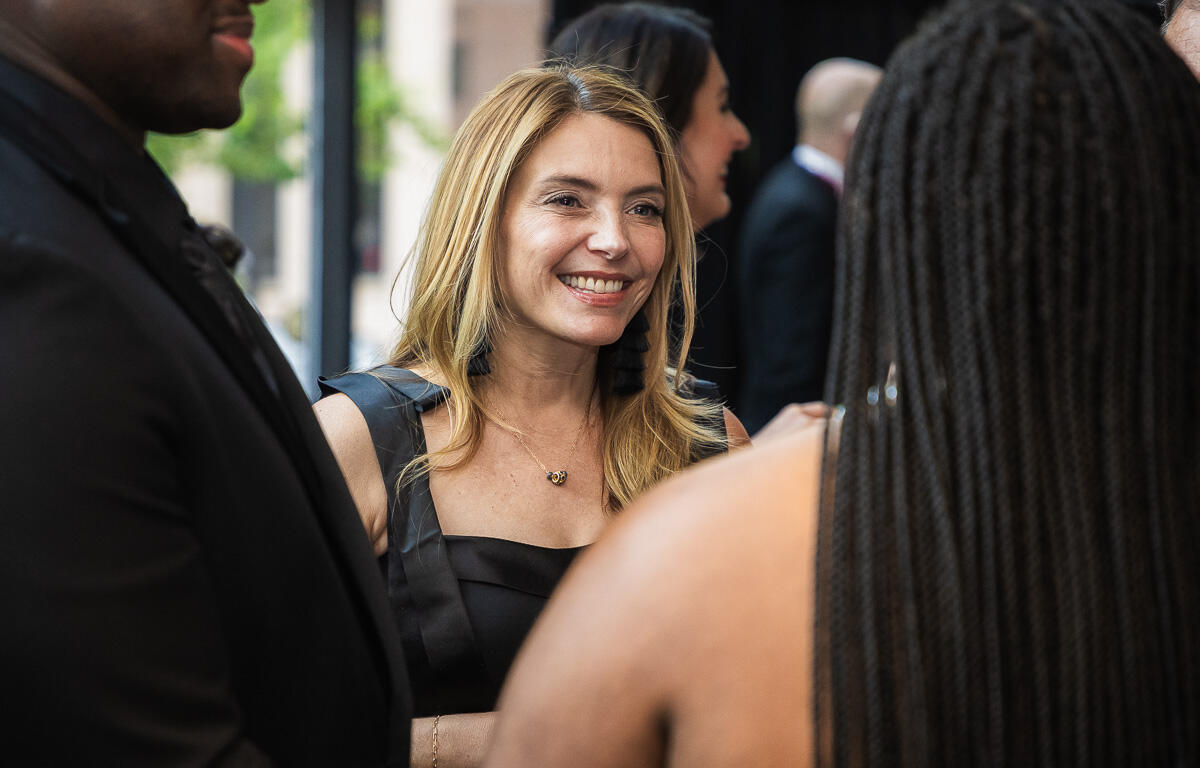 Smiling blonde woman in a black dress chats with guests at a social event or reception around her.