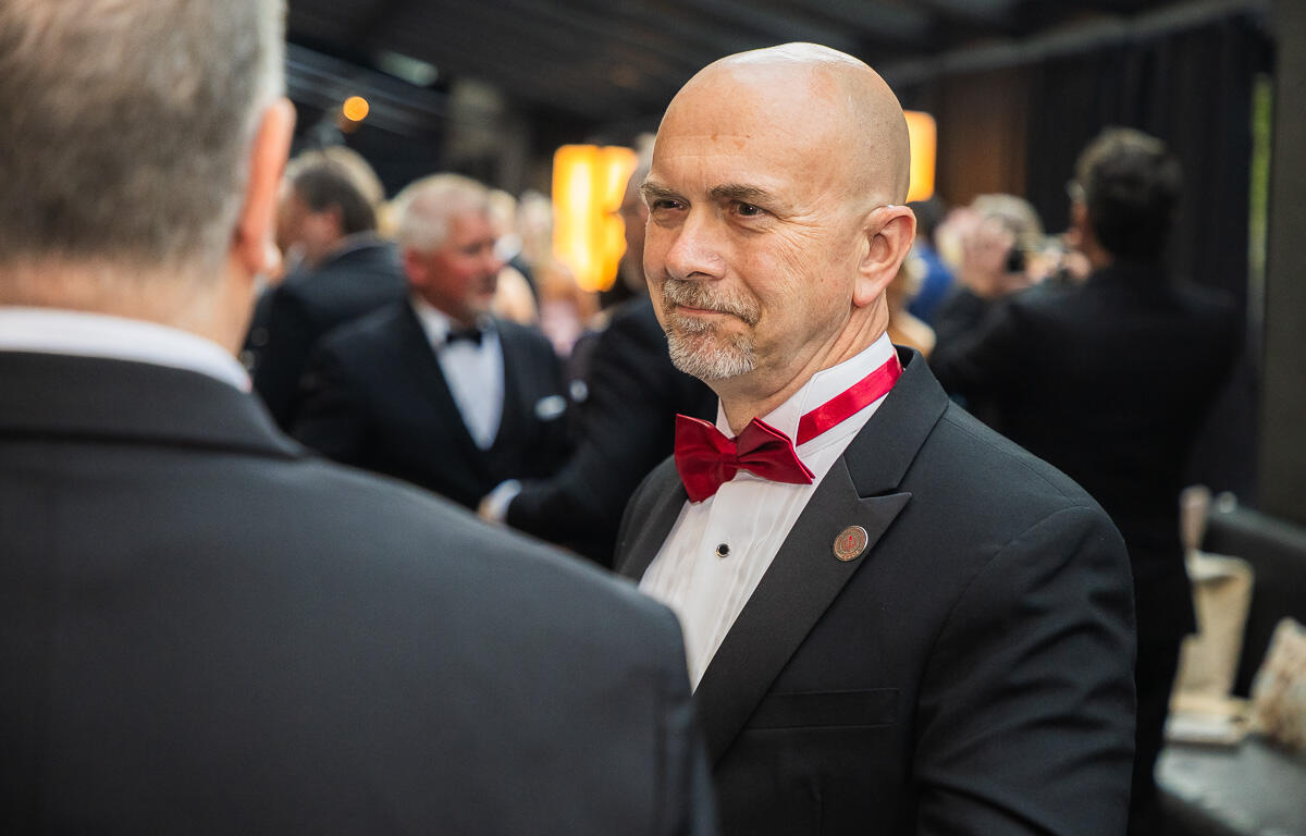 Bald man in a black tuxedo with a red bow tie at a formal event, conversing with another guest.
