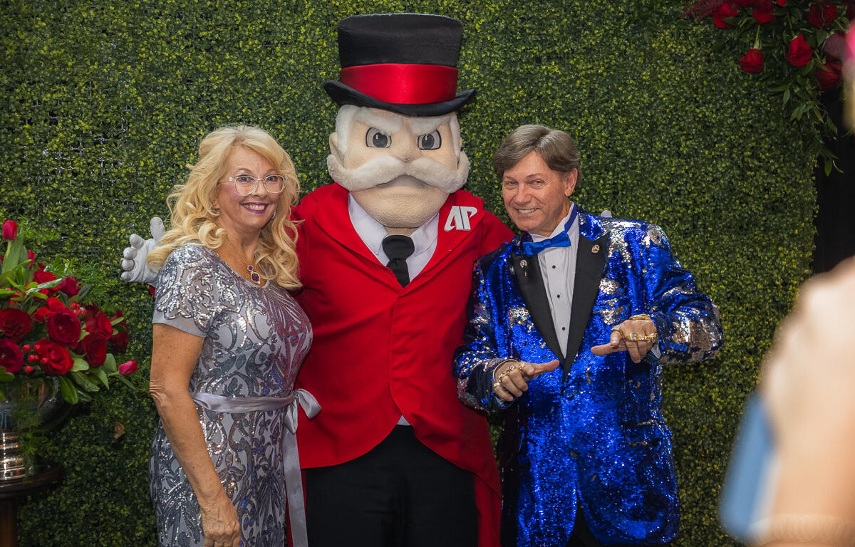 Woman in a silver sequined gown, a man in a blue glittery suit, and a mascot in a red jacket with a top hat posing together before a green hedge backdrop at a formal event.