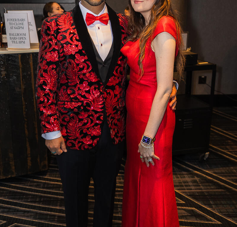 Couple posing together at a formal event; man in a red sequined jacket and bow tie, woman in a red gown, both smiling.