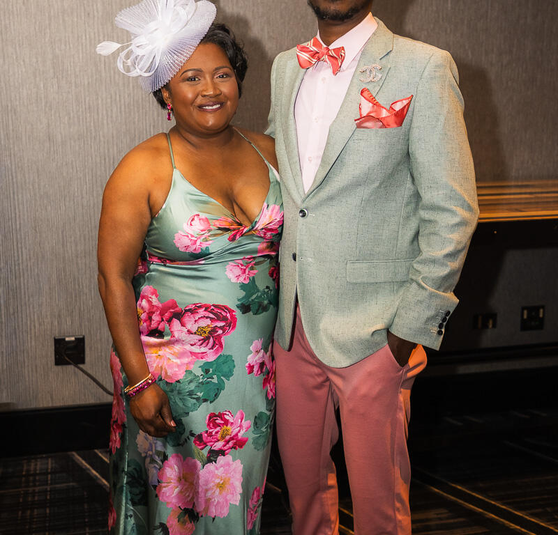 Couple posing at a formal event: woman in teal floral dress with a large white fascinator, man in pastel mint blazer, pink bow tie and pink pants with sunglasses.