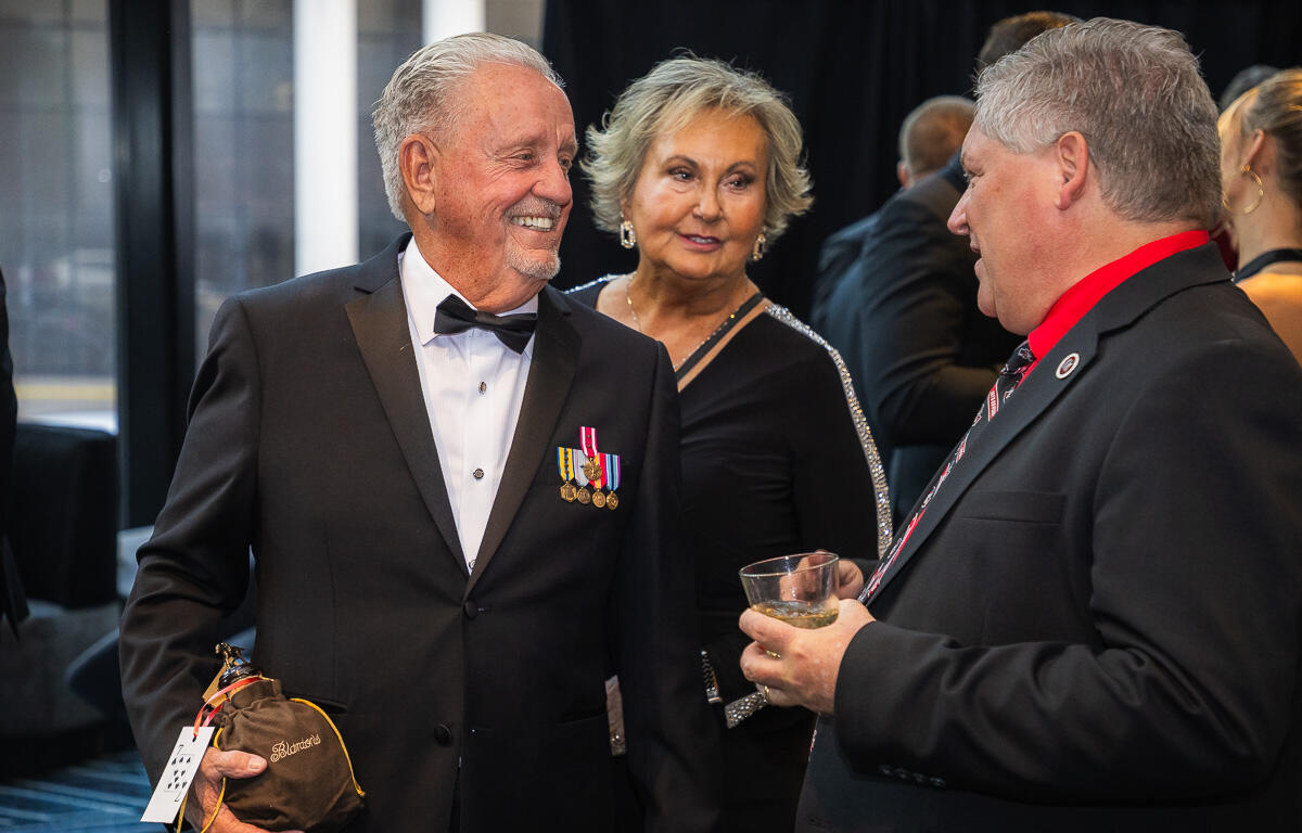 Older man in a tuxedo with medals smiles as he holds a wrapped gift bottle at a formal event; others chat in background.