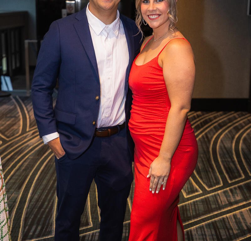 Couple posing together at a formal event: man in a navy suit and white shirt, woman in a red evening gown smiling at the camera, standing on a patterned carpet.