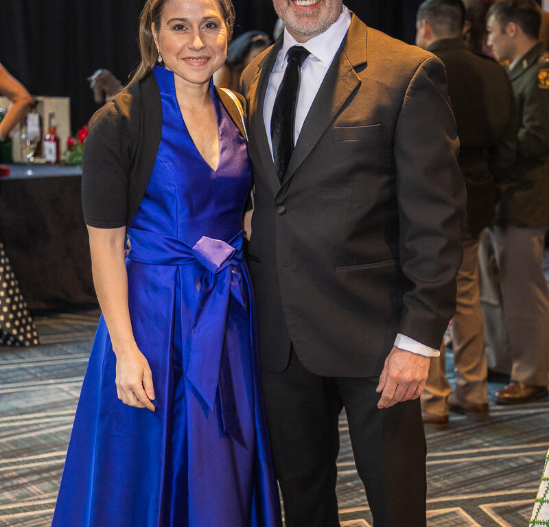 A smiling couple in formal attire posing together at a social event; woman in a blue gown and the man in a black suit and tie.