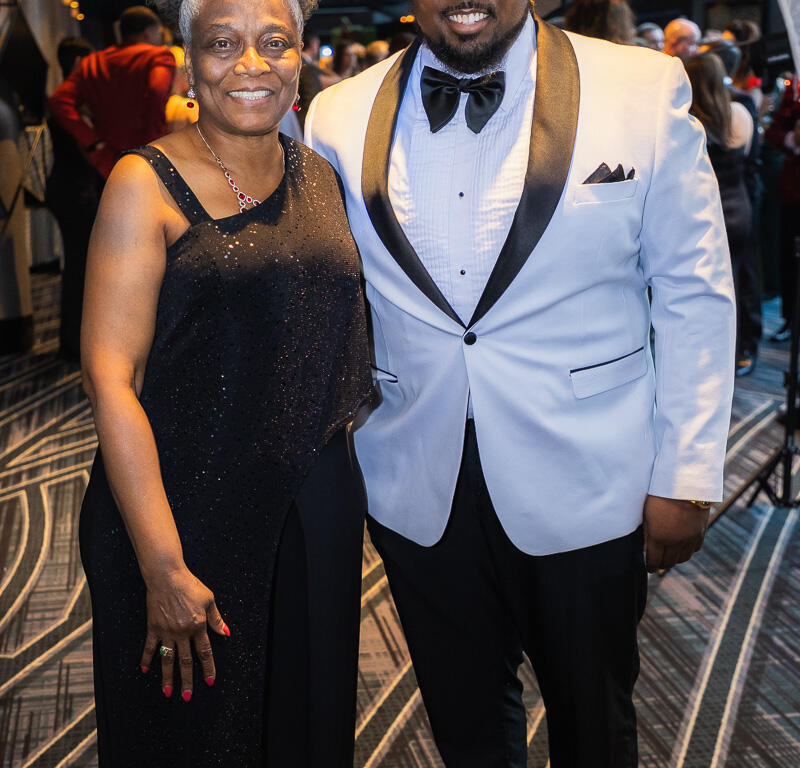 Smiling woman in a shimmering black evening gown beside a man in a white tuxedo jacket at a formal event.