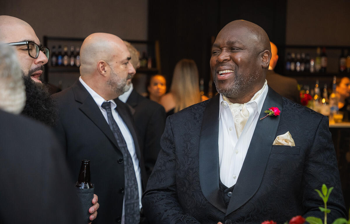 Man in a patterned tuxedo with cream vest and boutonniere smiling at a social event, bar in background.