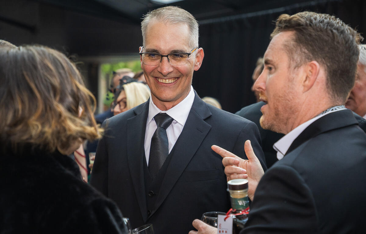 Smiling man in a dark suit and glasses chats at a formal gathering, holding a drink in a crowded event setting.