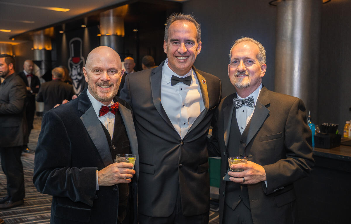 Three smiling men in tuxedos pose together at a formal event, each holding a drink in a dimly lit venue.