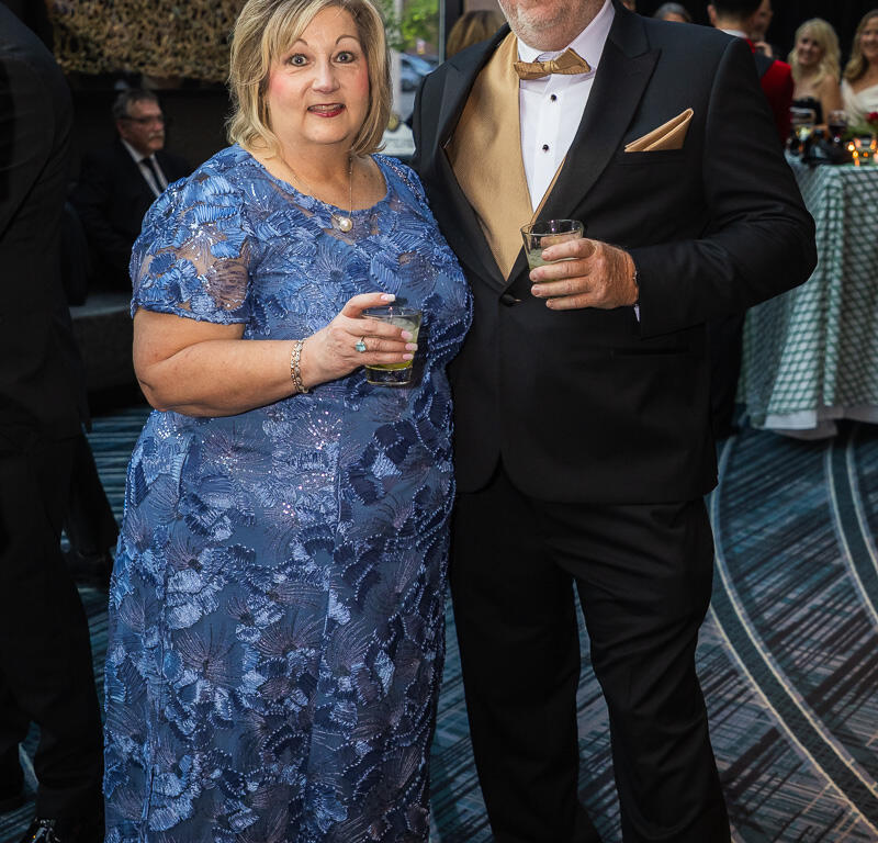 Couple at a formal event: woman in a blue lace evening gown with jewelry and a man in a black tuxedo with a gold vest and bow tie, each holding a drink.