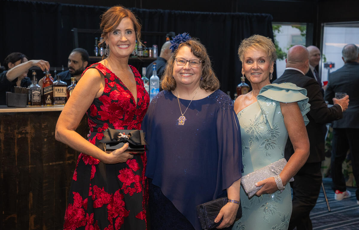 Three women in formal dresses pose together at a bar during a gala, each holding a clutch purse.